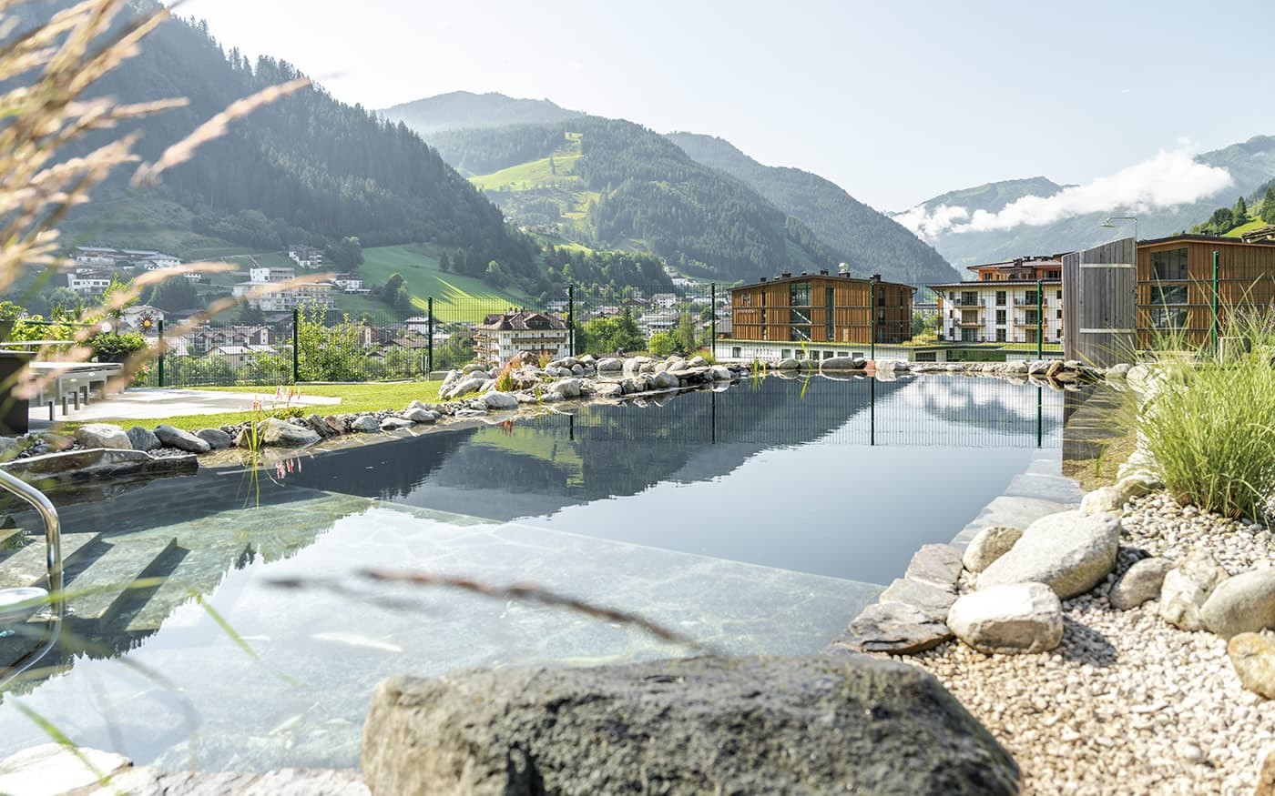 Ein Pool mit Steinen und Pflanzen vor einer Bergkulisse und modernen Gebäuden