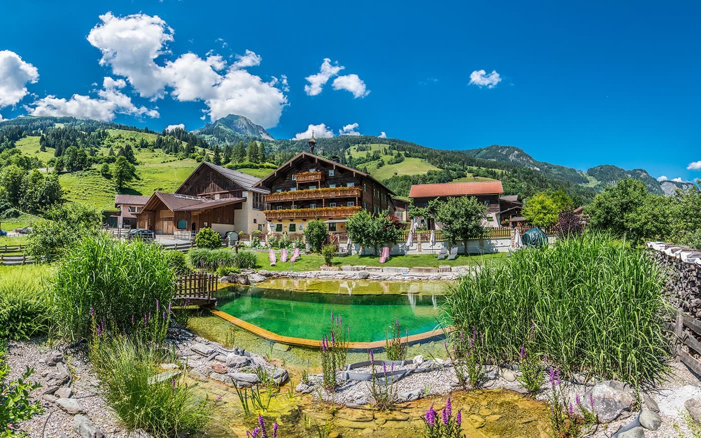 Ein malerisches Bauernhaus vor grünen Bergen und blauem Himmel mit einem kleinen Teich im Vordergrund